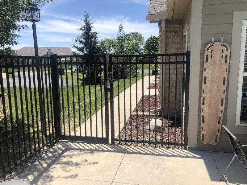 Black metal gate at a sunny residential patio opening to a concrete walkway and a grassy lawn beyond the fence.