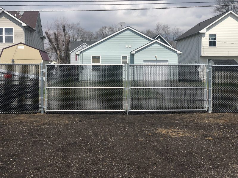 Metal chain-link fence with a double-swing gate in front of a row of pastel-colored houses in a suburban neighborhood.
